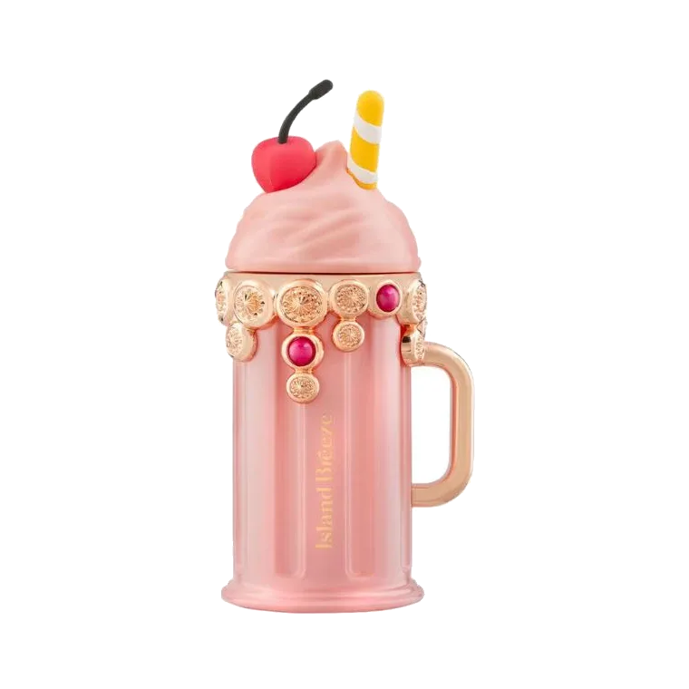 Frasco con forma de vaso de batido en vidrio rosa, con asa dorada, crema esculpida, una cereza, pajita a rayas y una guirnalda de pedrería dorada, sobre fondo blanco.